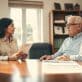 A serene office setting featuring a professional businesswoman in modest attire, discussing VA loan eligibility with an older gentleman, both appearing engaged and thoughtful. The foreground showcases a polished wooden desk with a VA loan pamphlet prominently displayed. In the middle ground, a window reveals a pleasant outdoor scene with soft, natural light streaming in, illuminating the room and creating a welcoming atmosphere. The background includes a bookshelf filled with documents and awards related to veteran services. The overall mood is one of support and guidance, emphasizing the importance of eligibility for surviving spouses. The image uses warm tones for a comforting yet professional feel, focusing on clarity and intimacy in the composition. - va loan closing costs
